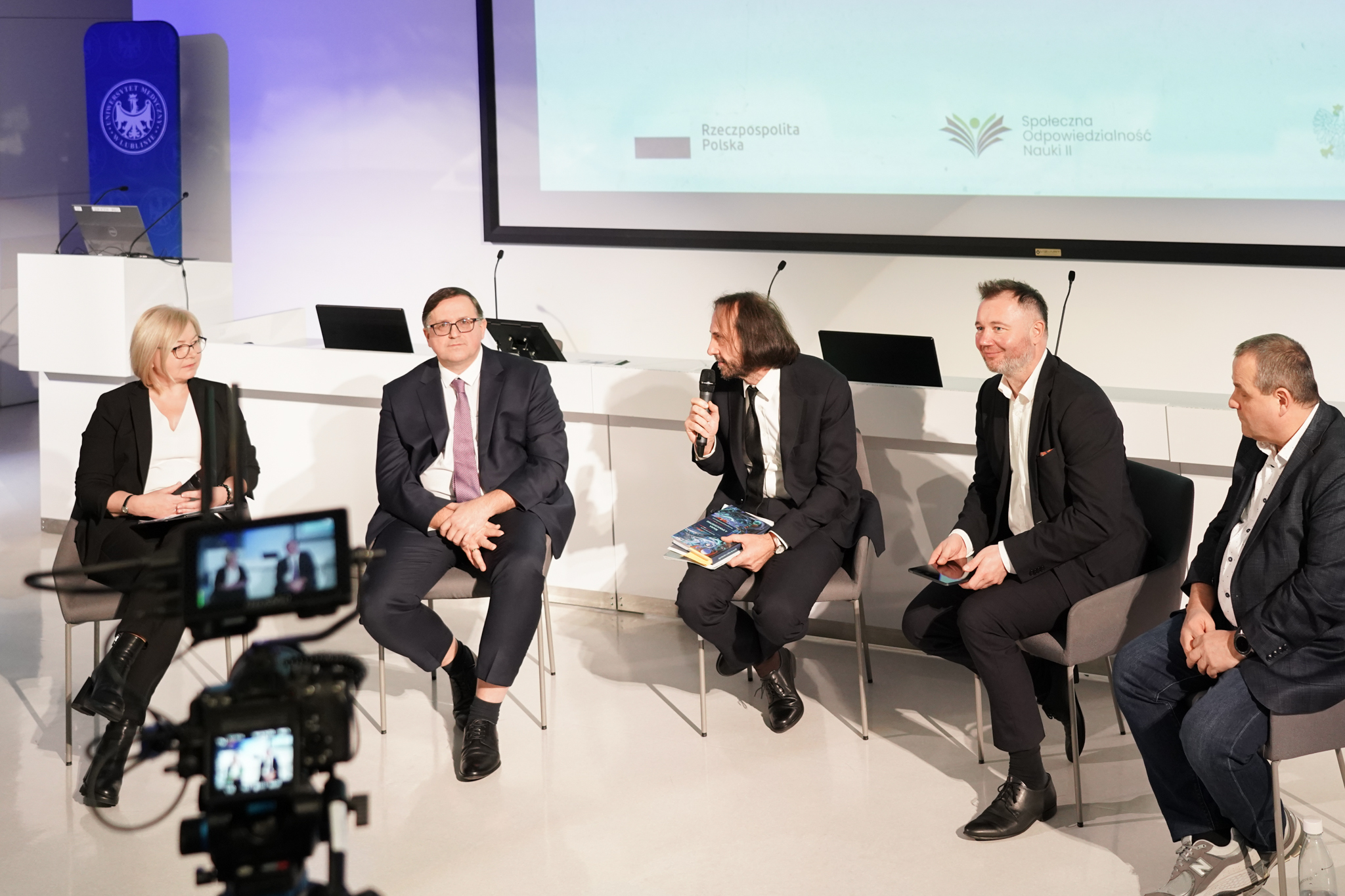 Pięć os&oacute;b w garniturach, to kobieta i czterech mężczyzn, mężczyzna w środku trzyma mikrofon, siedzą w sali wykładowej na scenie, z tyłu prezentacja wyświetlana na ścianie i st&oacute;ł z mikrofonami i laptopami